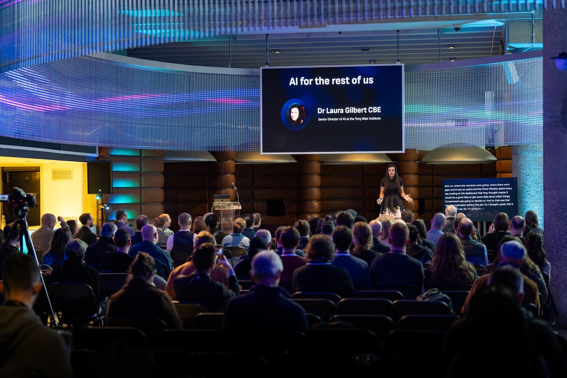 AI for the rest of us conference with live captioning by Procast AI - Dr Laura Gilbert CBE speaking to audience with live captions displayed on screen